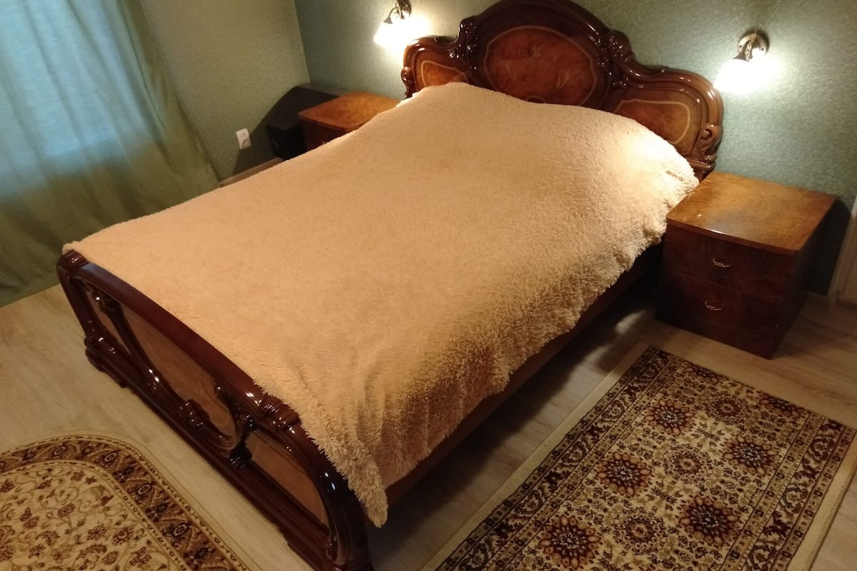 Een bed met houten frame en een witte deken, twee nachtkastjes met lampen, en een kamerkleed op de vloer.