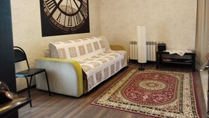 A living room with a sofa, a large wall clock, a radiator, and a patterned rug. - Apartments Galactika (Uralsk)