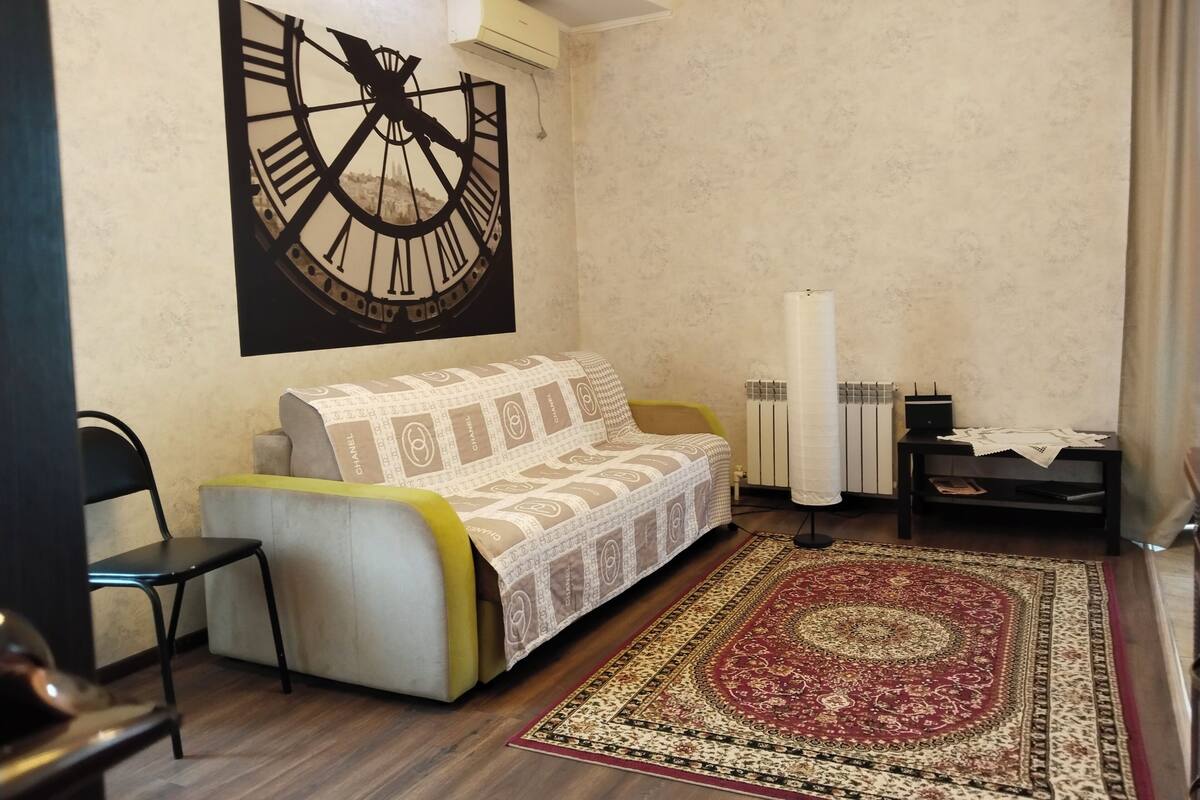 Een woonkamer met een bank, een grote wandklok, een radiator en een kamerbreed tapijt.