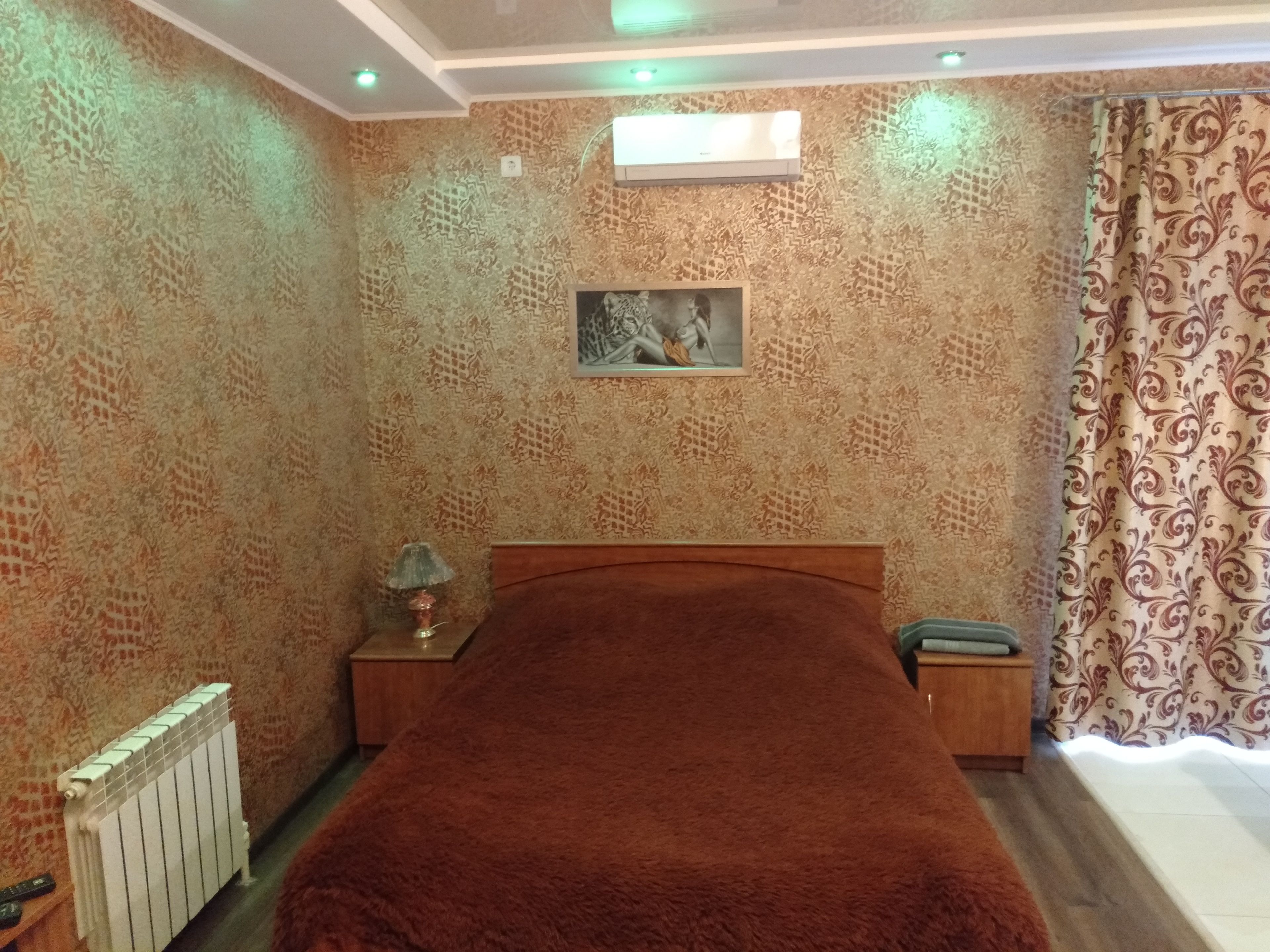 A bedroom with patterned wallpaper, a bed with a brown blanket, a nightstand with a lamp, a radiator, and a window with curtains.