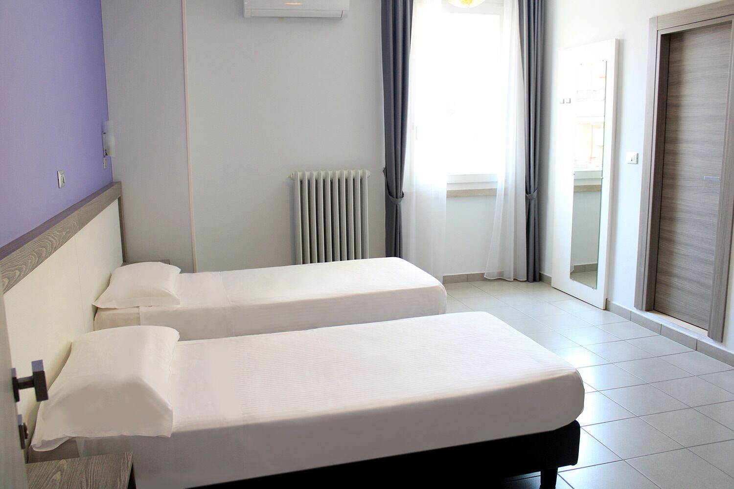 A hotel room with two single beds, a radiator, a window with curtains, and a sliding door.