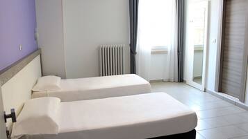 A hotel room with two single beds, a radiator, a window with curtains, and a sliding door.