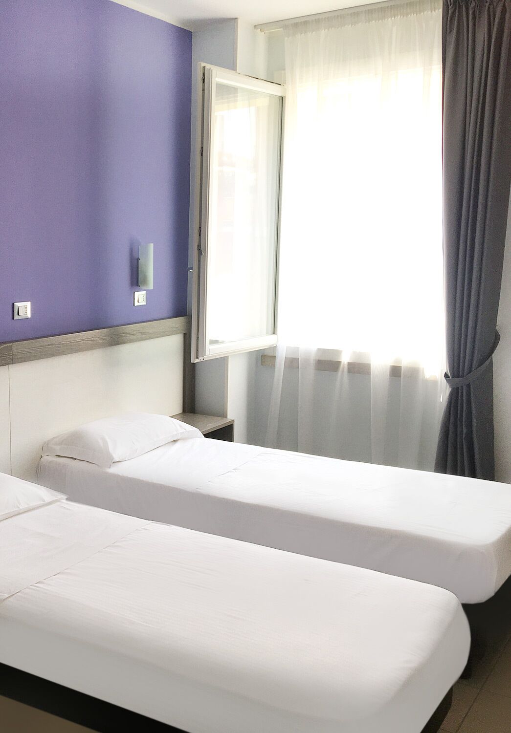A room with two single beds, a window with sheer curtains, and a wall-mounted light fixture.