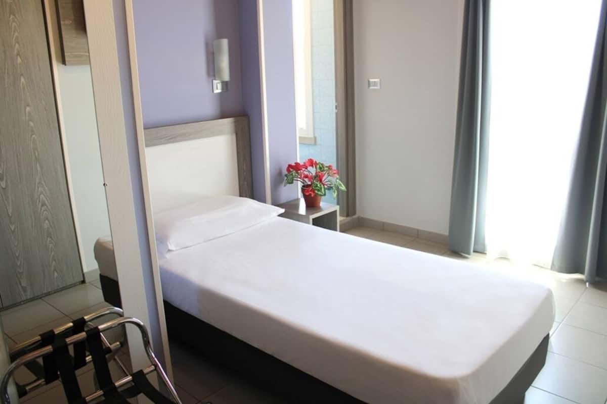 A single bed with a wooden headboard, a small bedside table with a potted plant, and a view of a window with curtains.