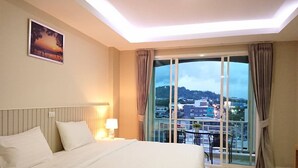 Une chambre d’hôtel avec un grand lit, une table de chevet avec une lampe, une vue sur la ville grâce à de grandes fenêtres et un tableau accroché au mur.