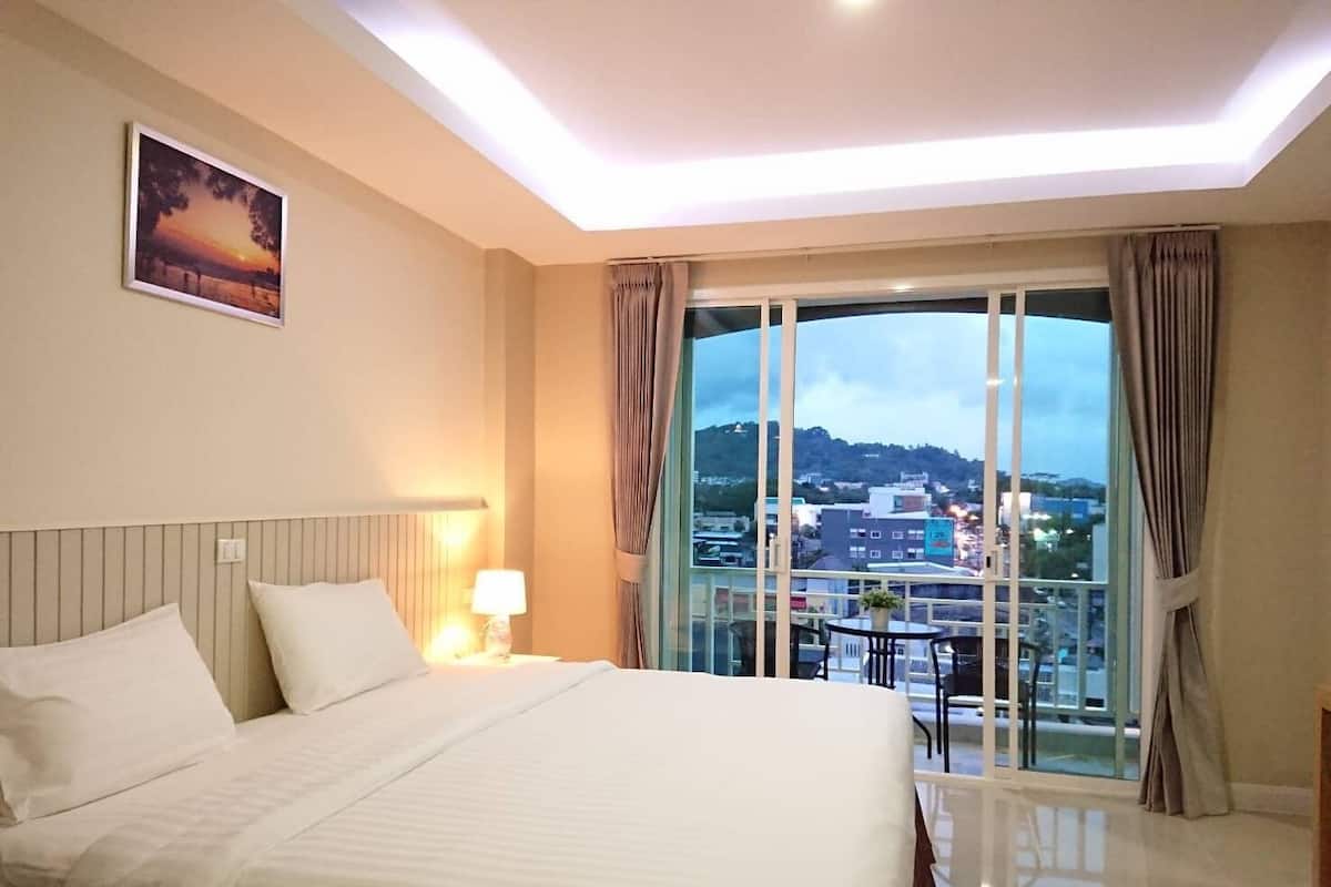 A hotel room with a large bed, a nightstand with a lamp, a view of the city through large windows, and a painting on the wall.