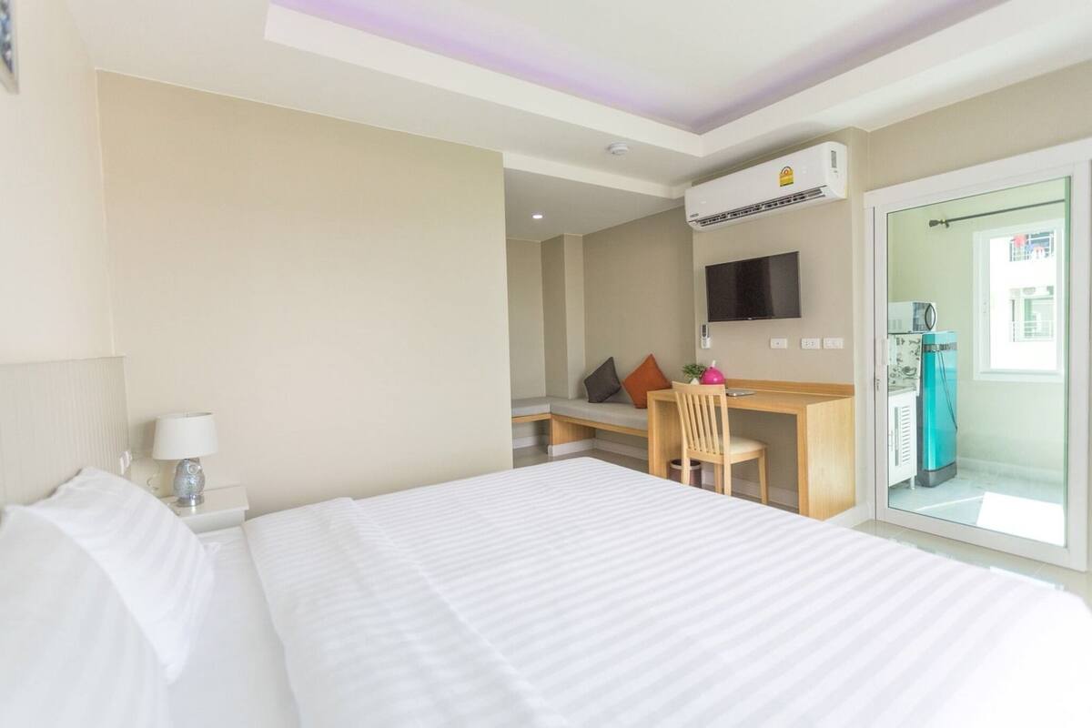 A hotel room with a bed, a desk, a chair, a TV, and an air conditioning unit.