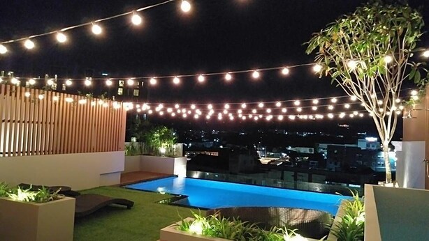 Un espace piscine aménagé sur le toit, avec des guirlandes lumineuses, une terrasse en bois et une vue sur le paysage urbain la nuit.