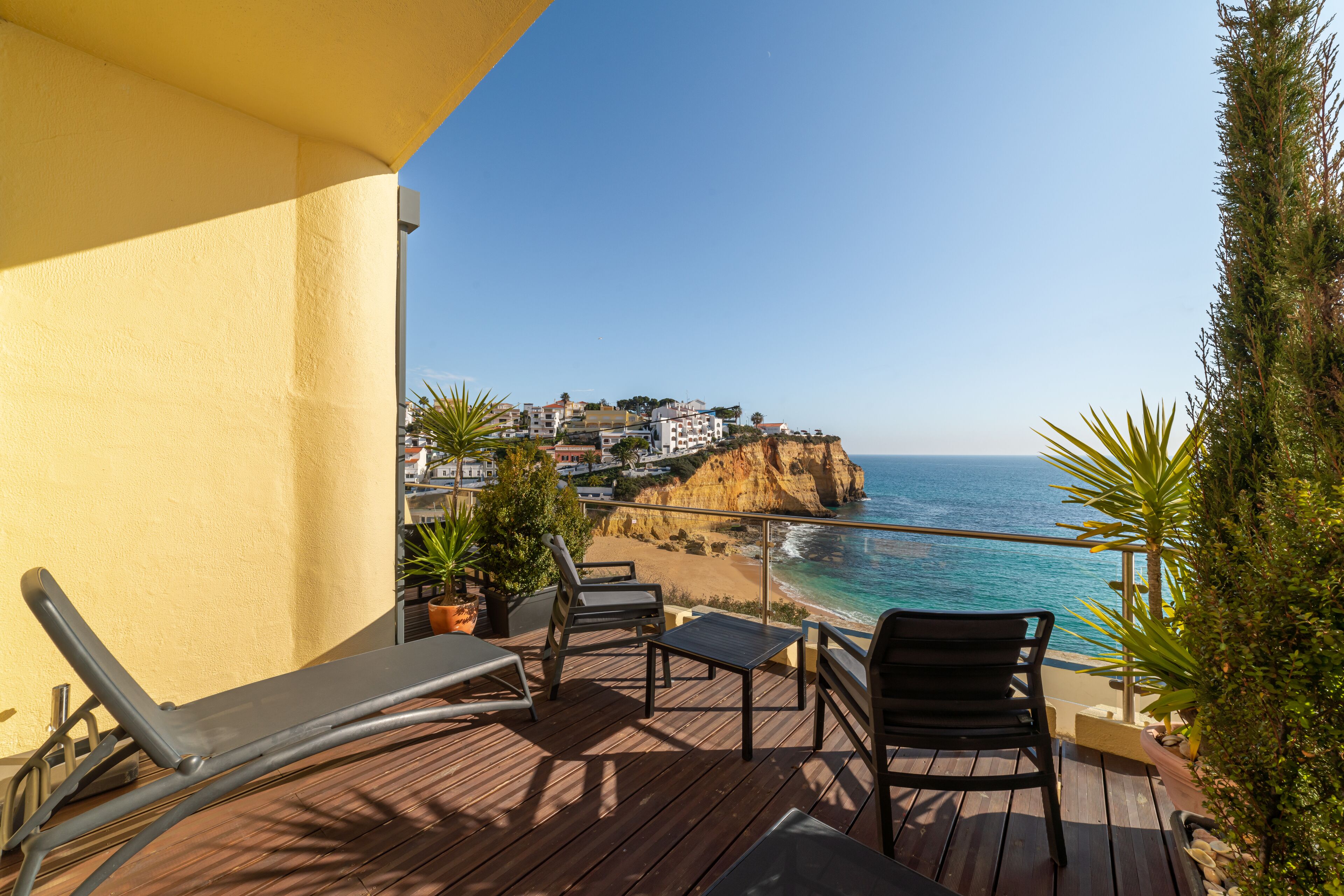 Terraza de madera con sillones de descanso y una mesa, con vistas a un pueblo costero y al mar.