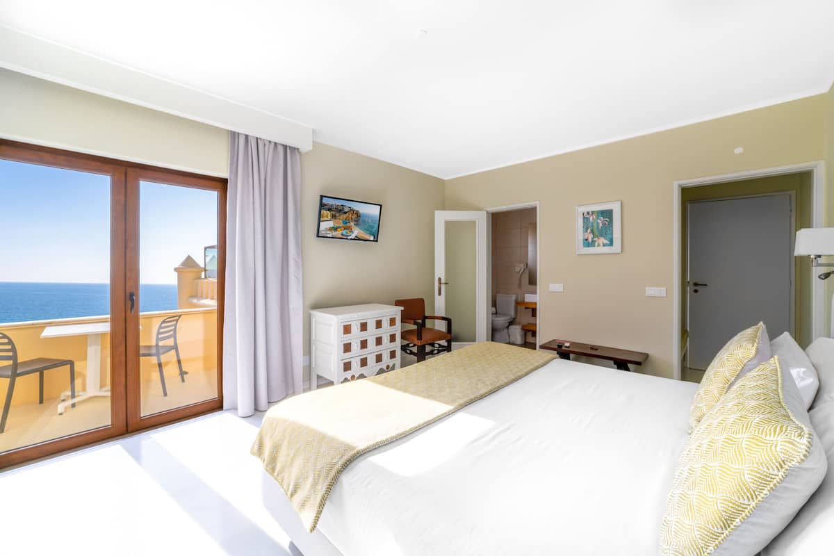A hotel room with a large bed, a desk, a chair, a small cabinet, and a balcony with a view of the sea.