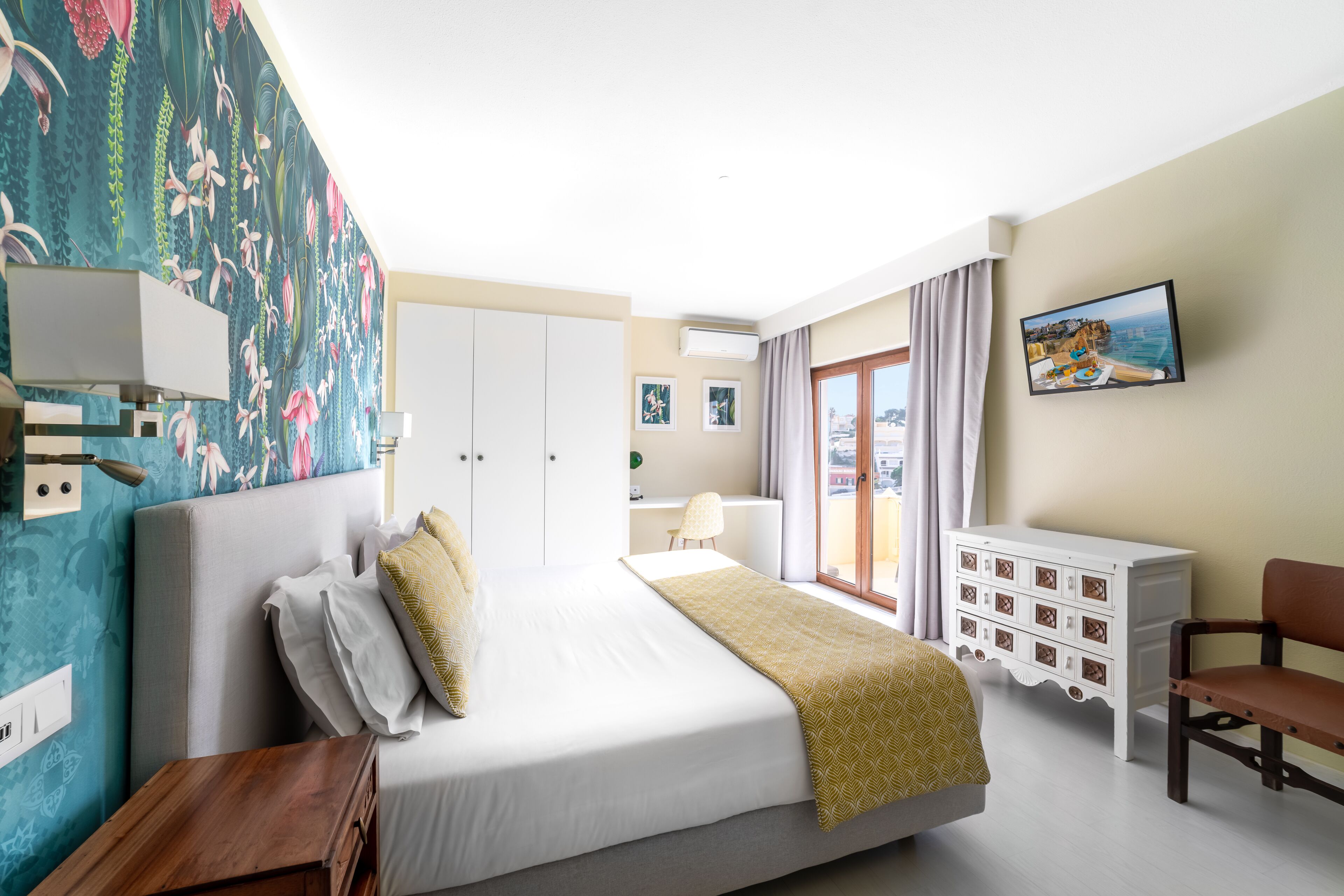 A hotel room with a bed, a desk, a chair, a dresser, and a large window with curtains.
