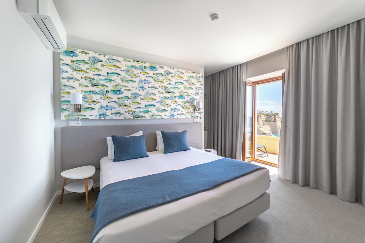 A modern hotel room with a large bed, a small table, a nightstand, a wall-mounted air conditioner, a ceiling light, and a view of the outdoors through a sliding glass door.