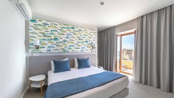 A modern hotel room with a large bed, a small table, a nightstand, a wall-mounted air conditioner, a ceiling light, and a view of the outdoors through a sliding glass door.