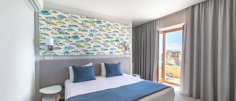 A modern hotel room with a large bed, a small table, a nightstand, a wall-mounted air conditioner, a ceiling light, and a view of the outdoors through a sliding glass door.