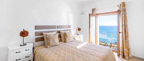 Un dormitorio con cama, mesita de noche, cómoda y vistas al mar a través de una puerta abierta.
