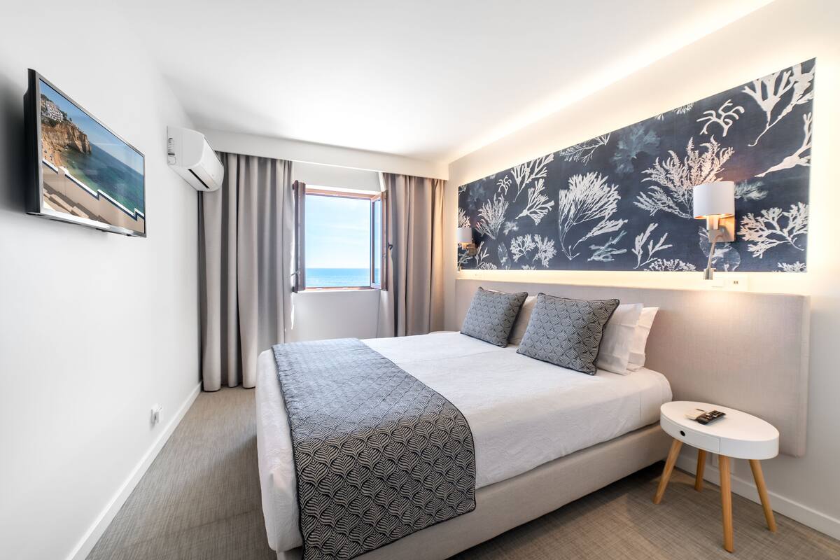 A modern hotel room with a large bed, a side table, a wall-mounted TV, and a view of the sea through the window.