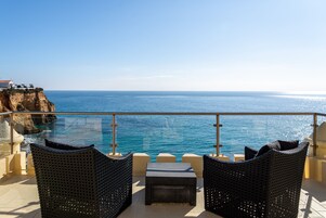 A balcony with wicker furniture overlooking a clear blue sea and a rocky coastline.