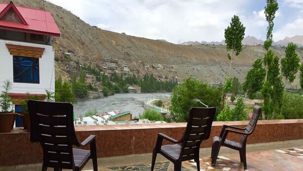 Una terraza con sillas que da a un río, un edificio de techo rojo y un paisaje montañoso al fondo.