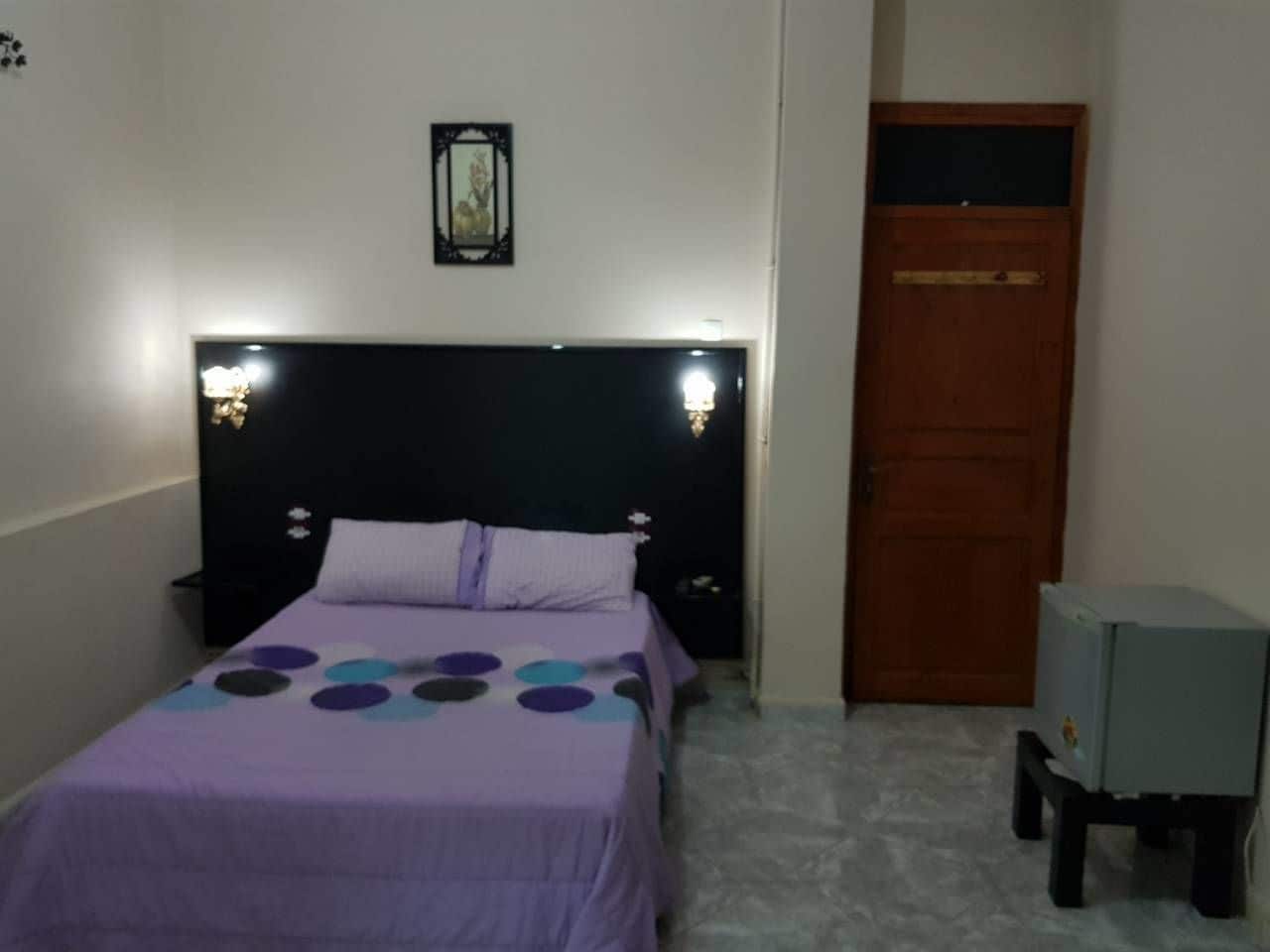 Una cama individual con cabecera, una mesita de noche, una lámpara de pared, una puerta y un televisor pequeño sobre un mueble.