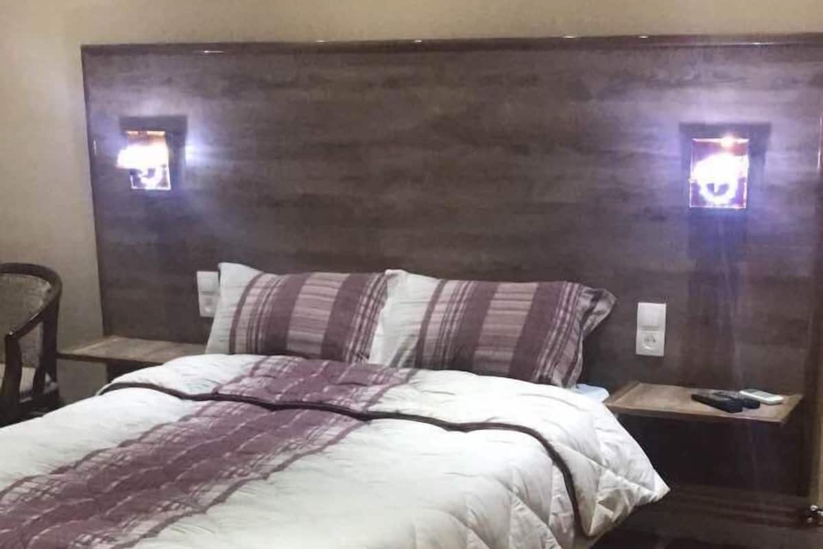 Cama bem arrumada com edredom acolchoado e almofadas, cabeceira de madeira e duas luminárias de mesa.