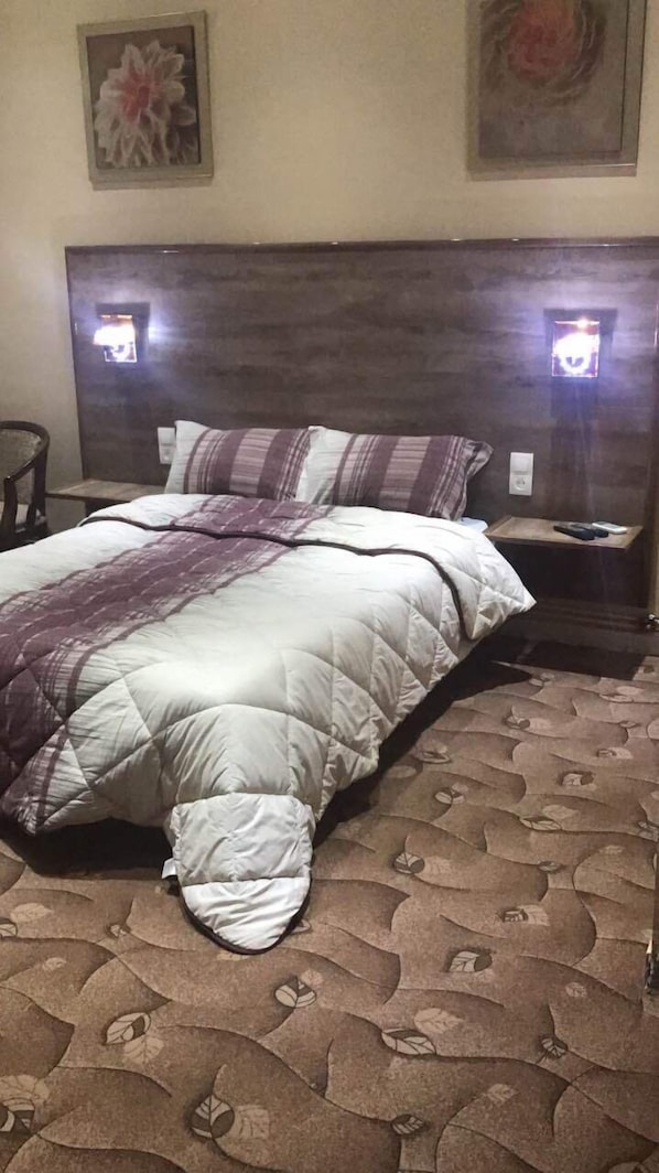 A neatly made bed with a quilted comforter and pillows, a wooden headboard, and two bedside lamps. - Hotel Ikram Oran (Oran)