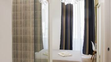 Een moderne hotelkamer met een glazen douchecabine, een bed met een witte dekbedovertrek en uitzicht op een raam met gestreepte gordijnen.