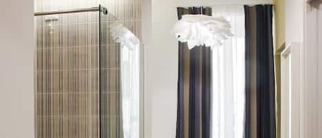 A modern hotel room with a glass shower enclosure, a bed with a white bedspread, and a view of a window with striped curtains.