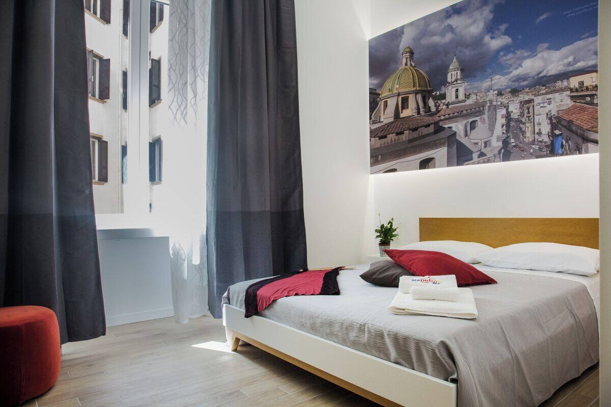 Une chambre d’hôtel avec un lit, un fauteuil rouge, un tableau représentant un paysage urbain et une fenêtre avec des rideaux.
