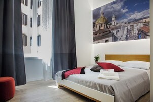 A hotel room with a bed, a red chair, a painting of a cityscape, and a window with curtains.