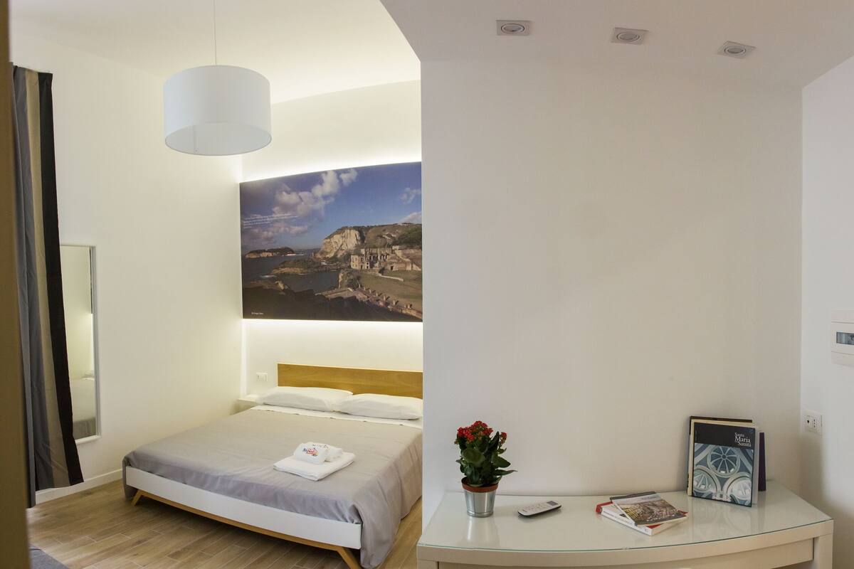 Une chambre d’hôtel avec un lit, un bureau, une plante en pot et un tableau représentant un paysage côtier.
