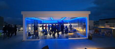 A modern glass-walled lounge area with seating and tables, illuminated by blue lighting.