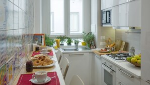 A modern kitchen with white cabinets, a stove, and a sink. There are various kitchen items on the counter, including a basket of fruit, a jar of jam, and a plate of pastries. - Napolinn B&B (Naples)