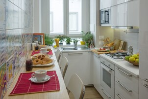 A modern kitchen with white cabinets, a stove, and a sink. There are various kitchen items on the counter, including a basket of fruit, a jar of jam, and a plate of pastries.