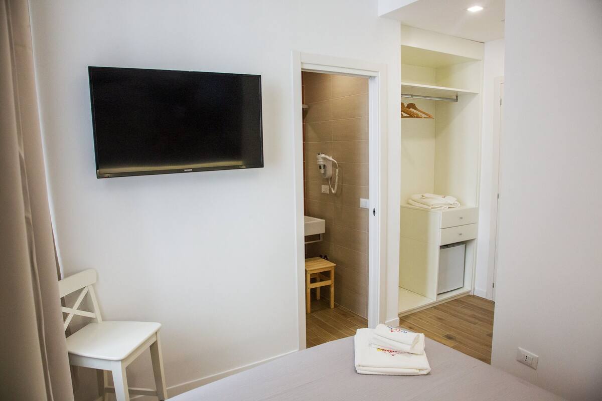 Une chambre d’hôtel équipée d’un téléviseur à écran plat fixé au mur, d’une chaise en bois, d’une petite table et d’une serviette soigneusement pliée posée sur le lit.