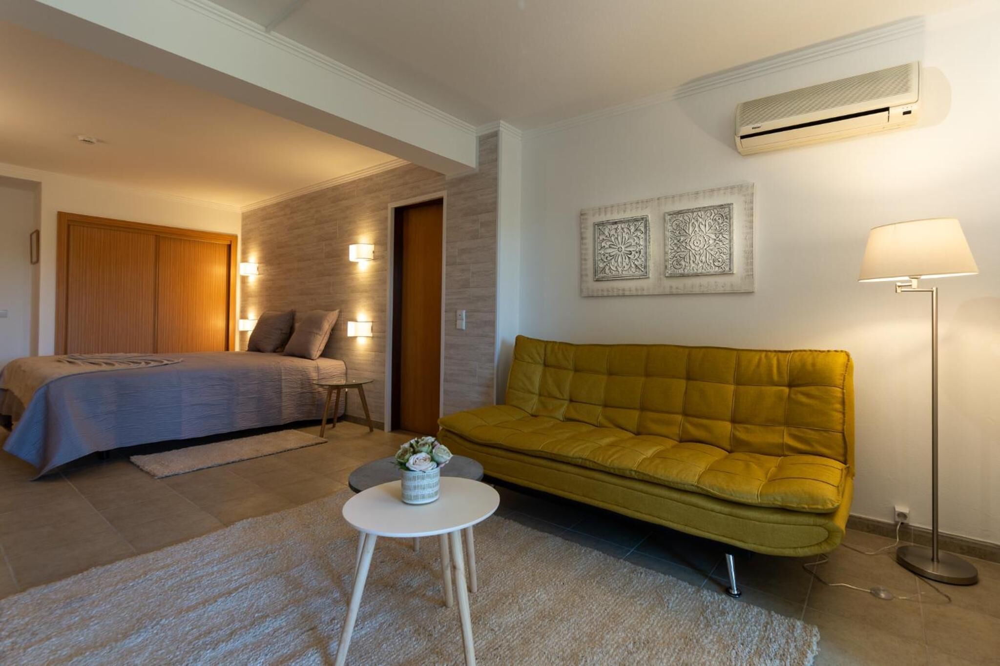 Un dormitorio moderno con una cama, un sofá, una mesita con un florero y un equipo de aire acondicionado.