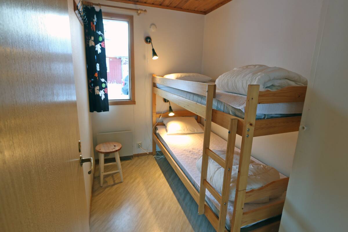 Ein kleiner, kompakter Raum mit Etagenbetten, einem kleinen Hocker und einem Fenster mit Vorhängen.