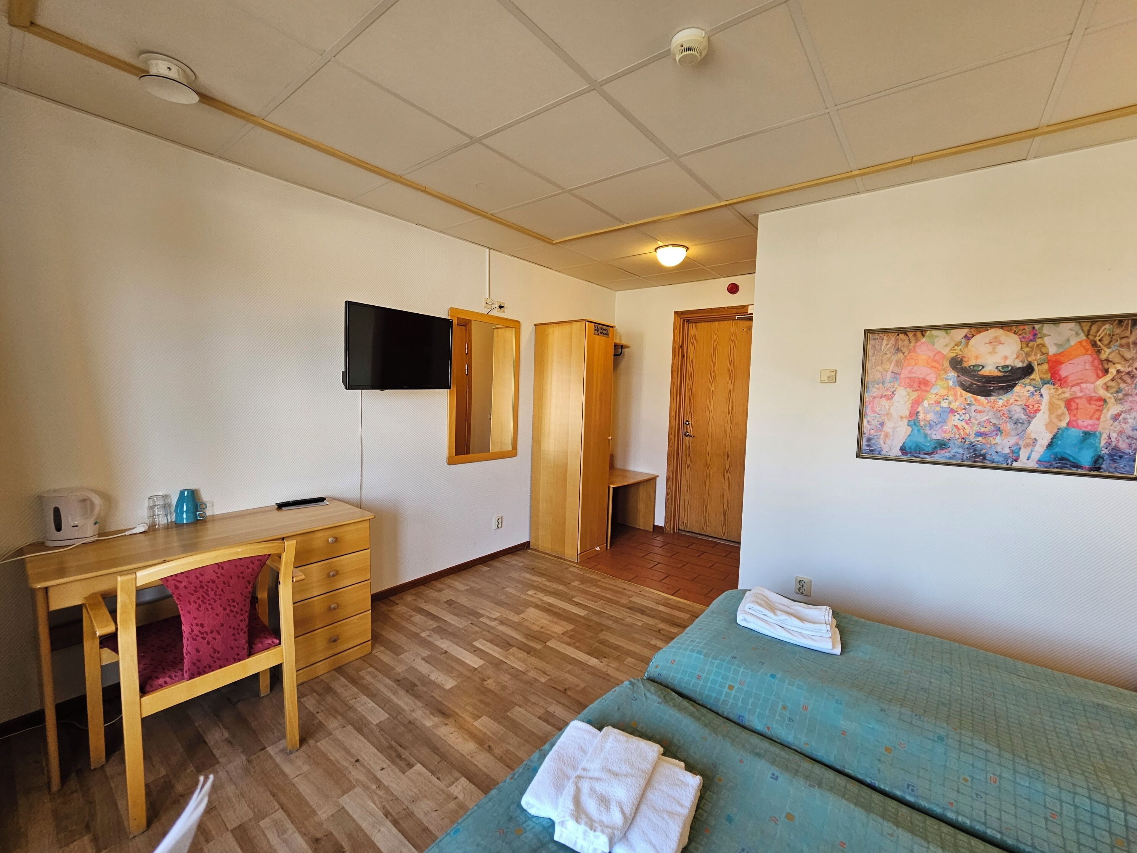 A room with a wooden desk, a television mounted on the wall, a bed with a blue mattress, and a wooden wardrobe.