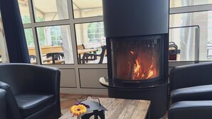 Una habitación acogedora con chimenea, sillones de cuero, una mesa de madera con un jarrón de flores y vistas al exterior a través de amplias ventanas.