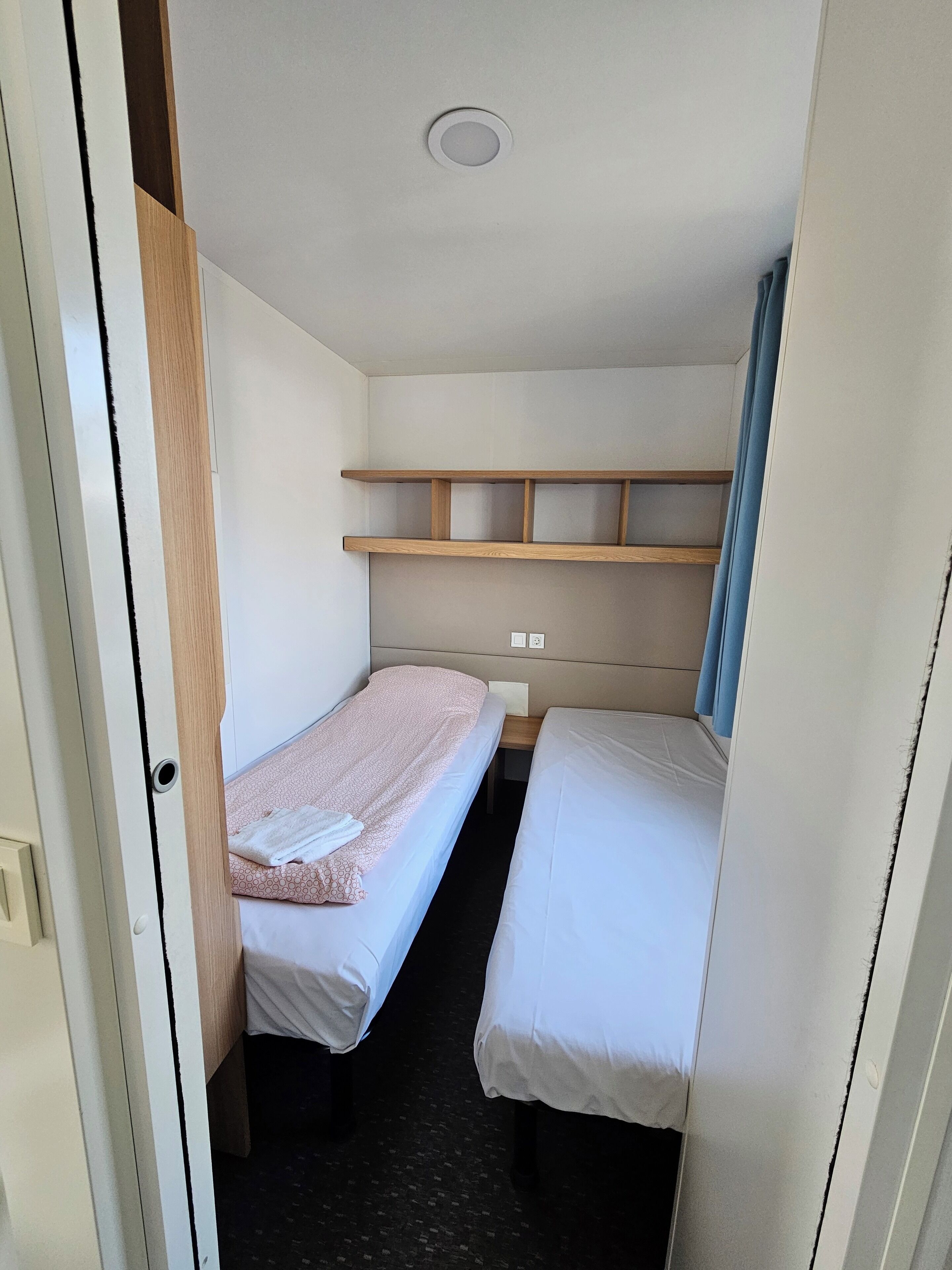 A compact room with two single beds, a wooden shelf, and a ceiling light.