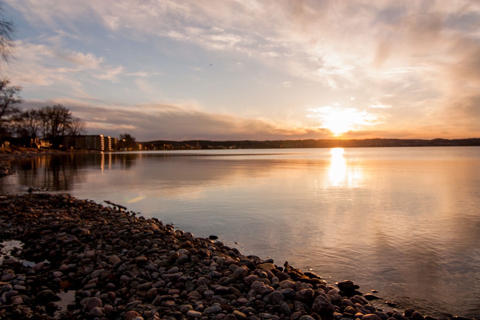 Un lago tranquilo al atardecer, con una orilla rocosa y edificios a lo lejos.