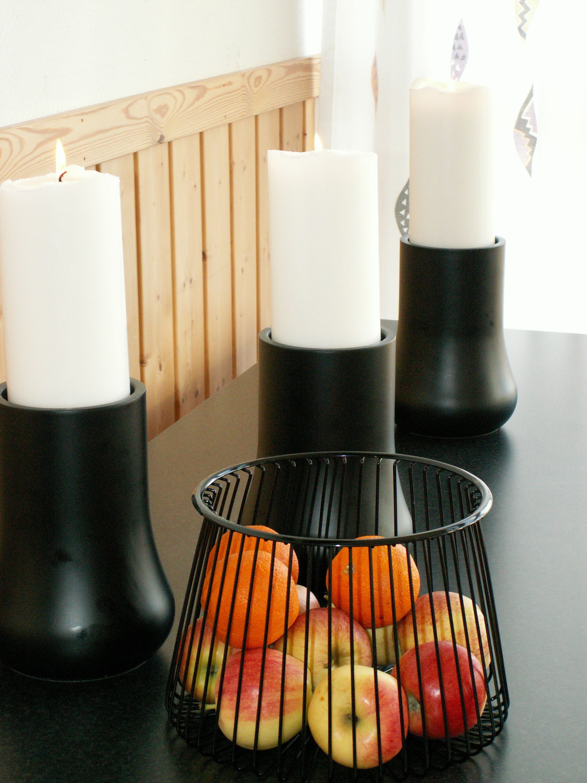 Three white candles in black holders on a table with a basket of apples and oranges.
