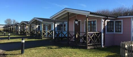 A row of pink mobile homes with wooden balconies, a paved walkway, and a grassy area.