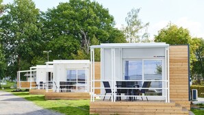 Una hilera de cabañas blancas y modernas de una sola habitación, con detalles de madera, cada una con balcón y un ventanal grande, ubicadas en un área cubierta de césped con árboles al fondo.