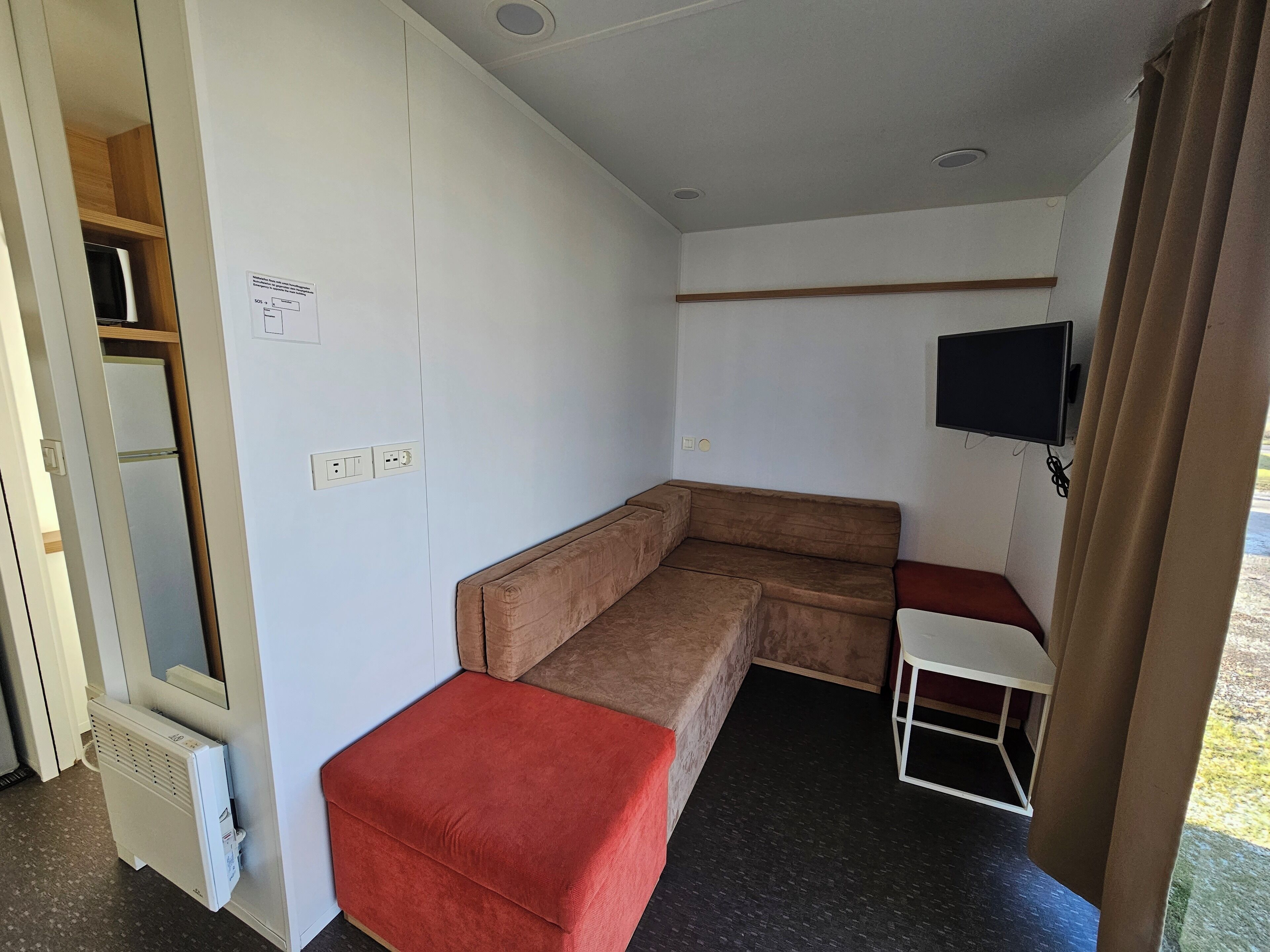 A compact room with a sofa, a small table, a television mounted on the wall, and a heater against the wall.