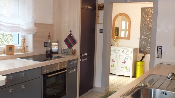 Una cocina moderna con horno empotrado, un lavamanos y un refrigerador. Hay una ventana con cortinas blancas y una puerta que conduce a otra habitación.