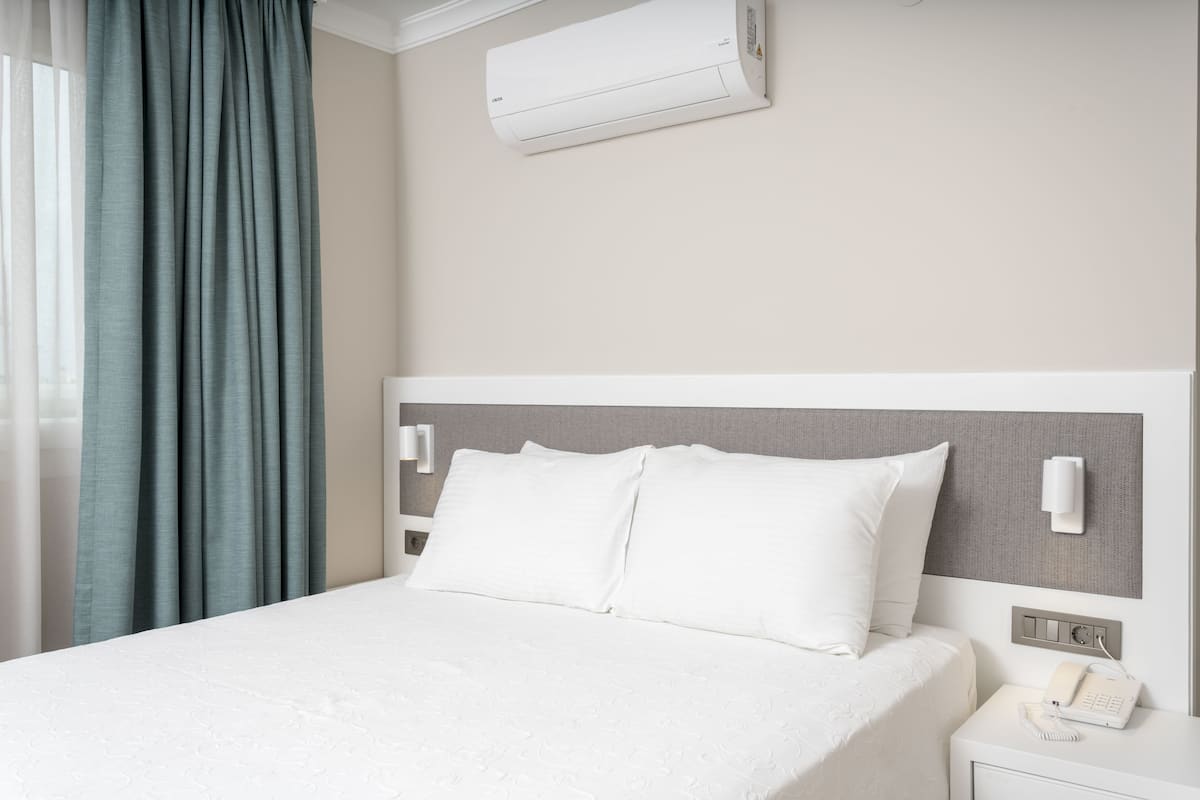 A neatly made bed with white linens, a bedside table with a phone, and a wall-mounted air conditioner.