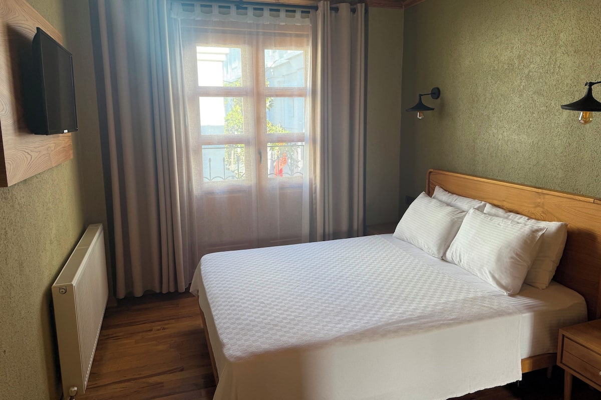 A bedroom with a bed, a nightstand, a wall-mounted TV, a window with curtains, and a radiator.