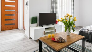 A modern hotel room with a wooden floor, a white TV stand with a flat-screen TV, a small plant, a vase with yellow flowers, a tray with pastries and glasses of orange juice, and a plaid blanket on a bed. - Green Garden Residence (Kraków)