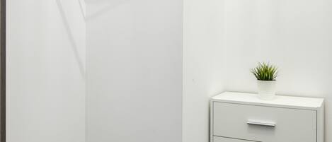 Een hoek van een kamer met een houten kledingrek, een klein wit kastje met lades, en een potplant erbovenop.