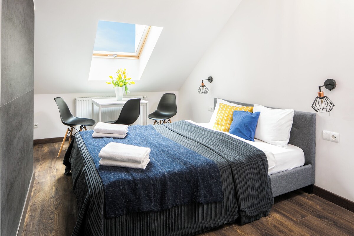 A modern bedroom with a bed, a desk, two chairs, a window, and a wall-mounted lamp.